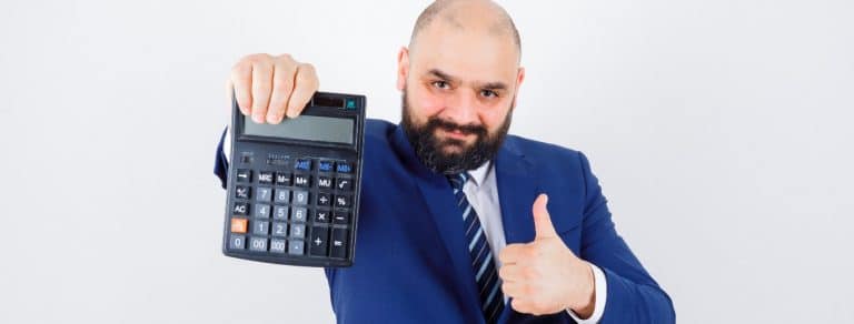 Homem branco, careca e com barba, faz um aceno de joia com a mão direita e segura uma calculadora na mão esquerda