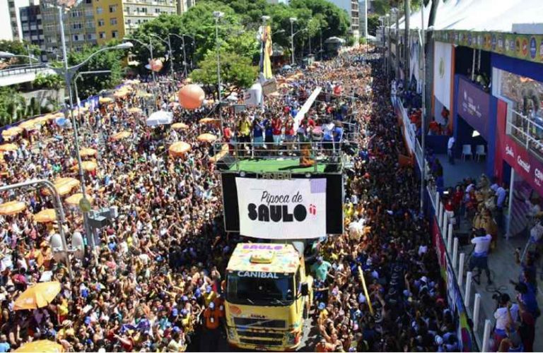 Multidão de pessoas em uma comemoração de rua, conhecido como bloco de carnaval de rua, com um trio elétrico cheio de artistas cantando para a população