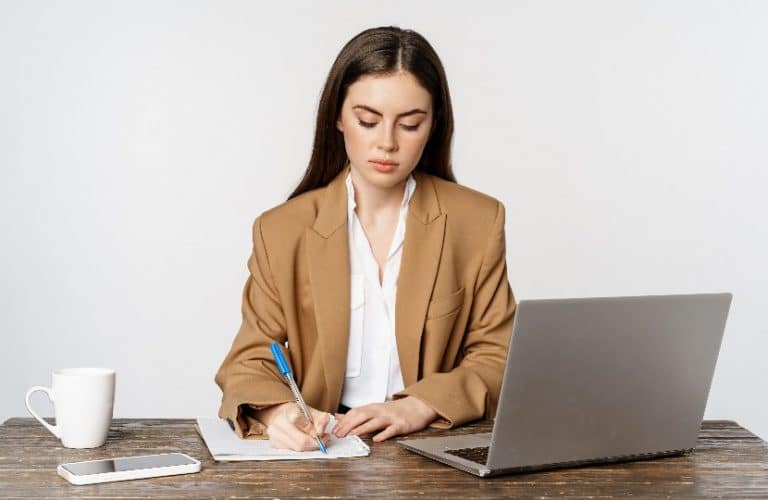 Mulher sentada em frente ao computador, anotando em um papel.