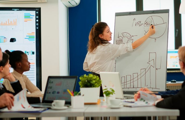 Mulher desenha no quadro em reunião, enquanto pessoas observam sentadas.