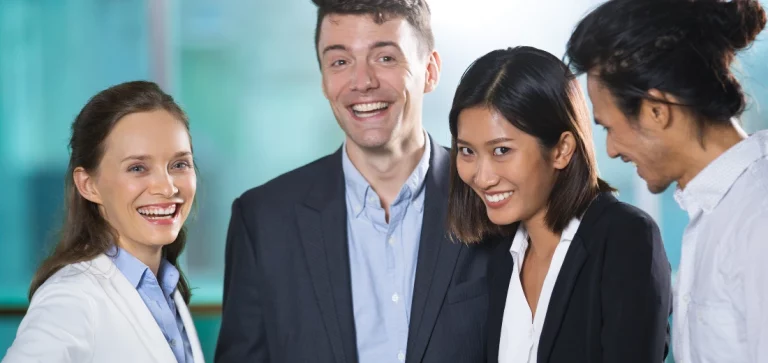 Mulher branca de olhos claros sorrindo, junto com um homem branco de terno sorrindo, uma mulher asiática olhando frontalmente e sorrindo, e um homem asiático olhando para a mulher asiática, sorrindo